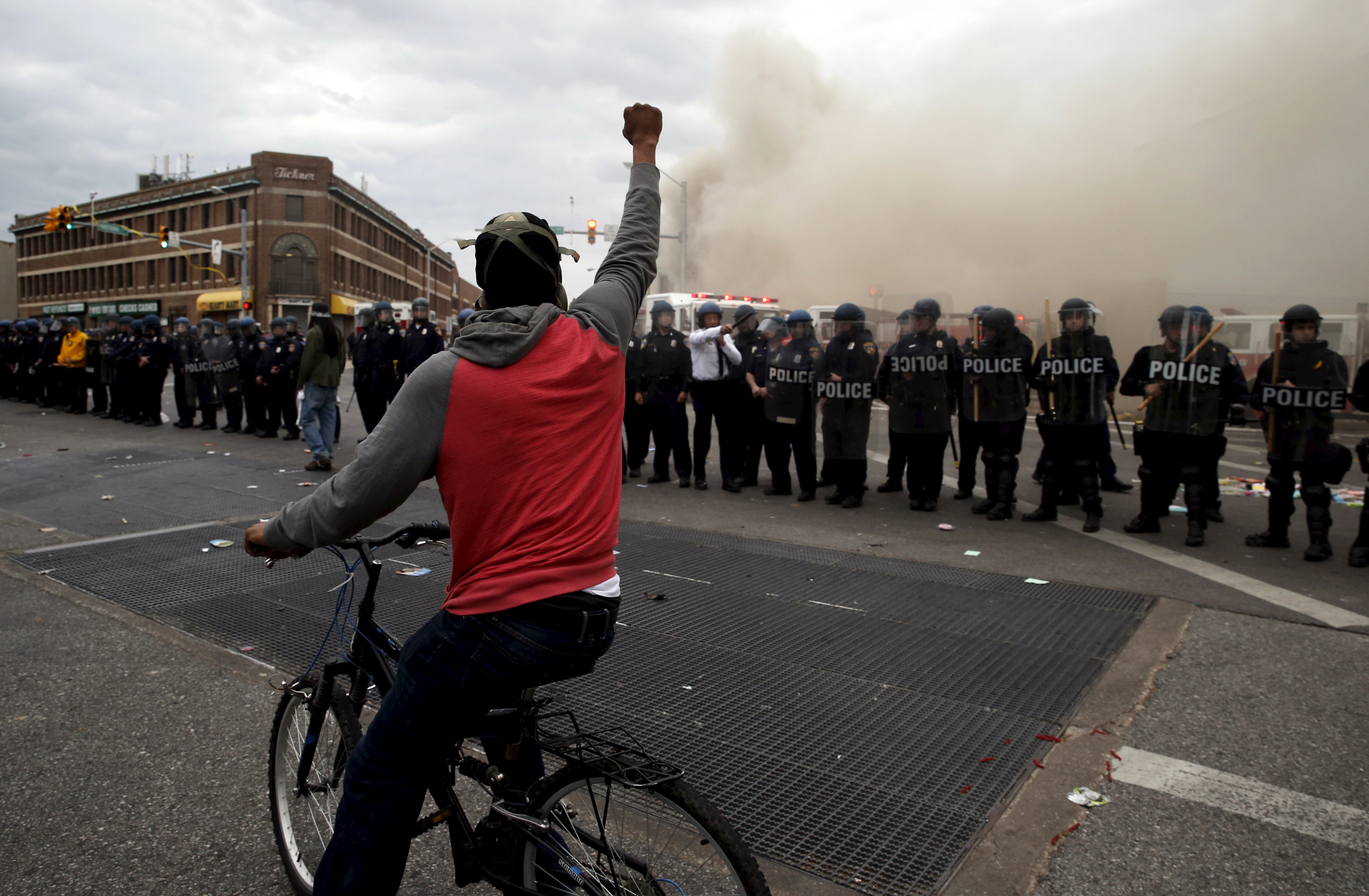 A protestor on a bicycle thrusts his fist in the air next to a line of police, in front of a burning CVS drug store, during clashes in Baltimore