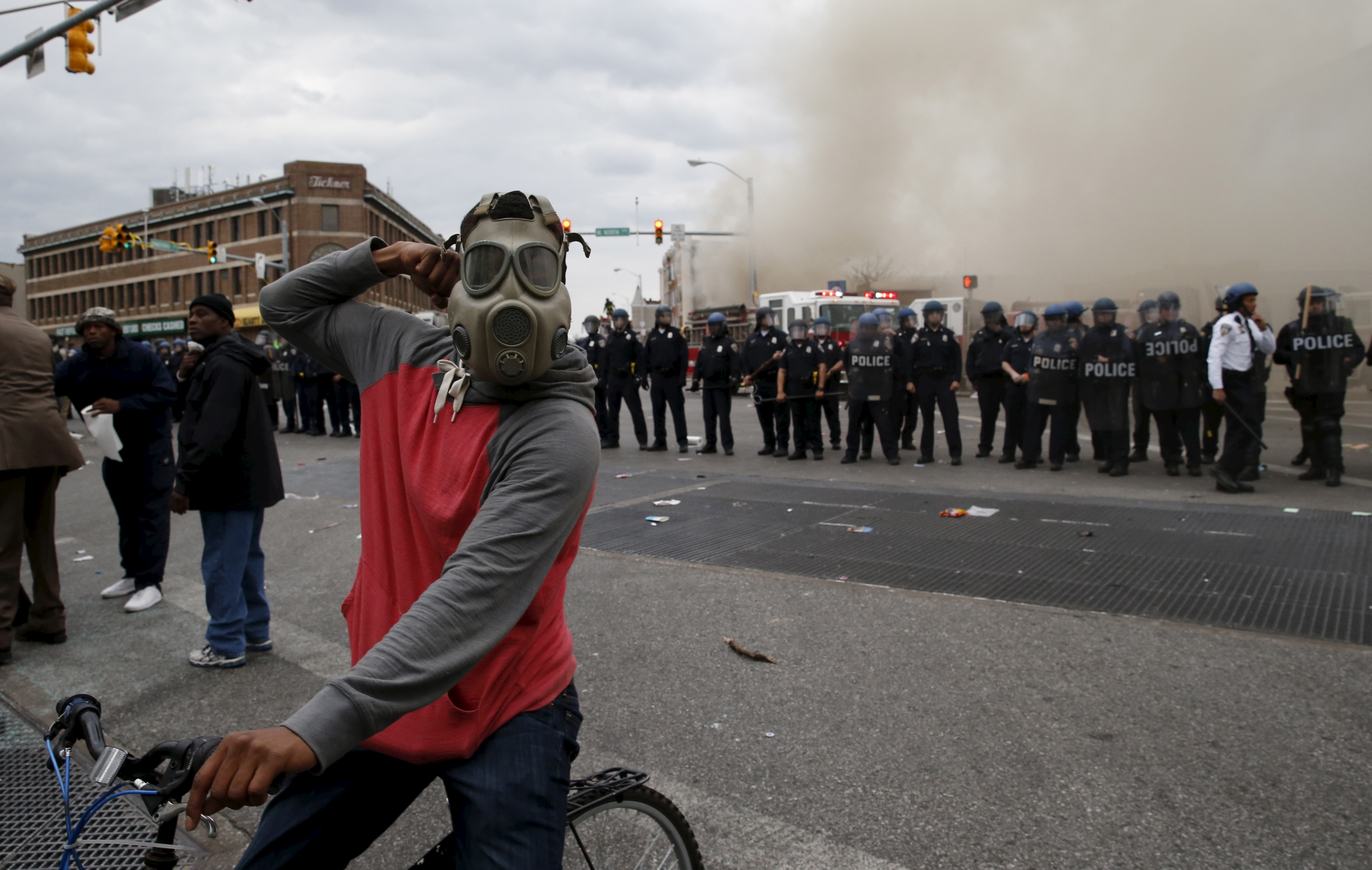 A protestor on a bicycle thrusts his fist in the air next to a line of police, in front of a burning CVS drug store, during clashes in Baltimore