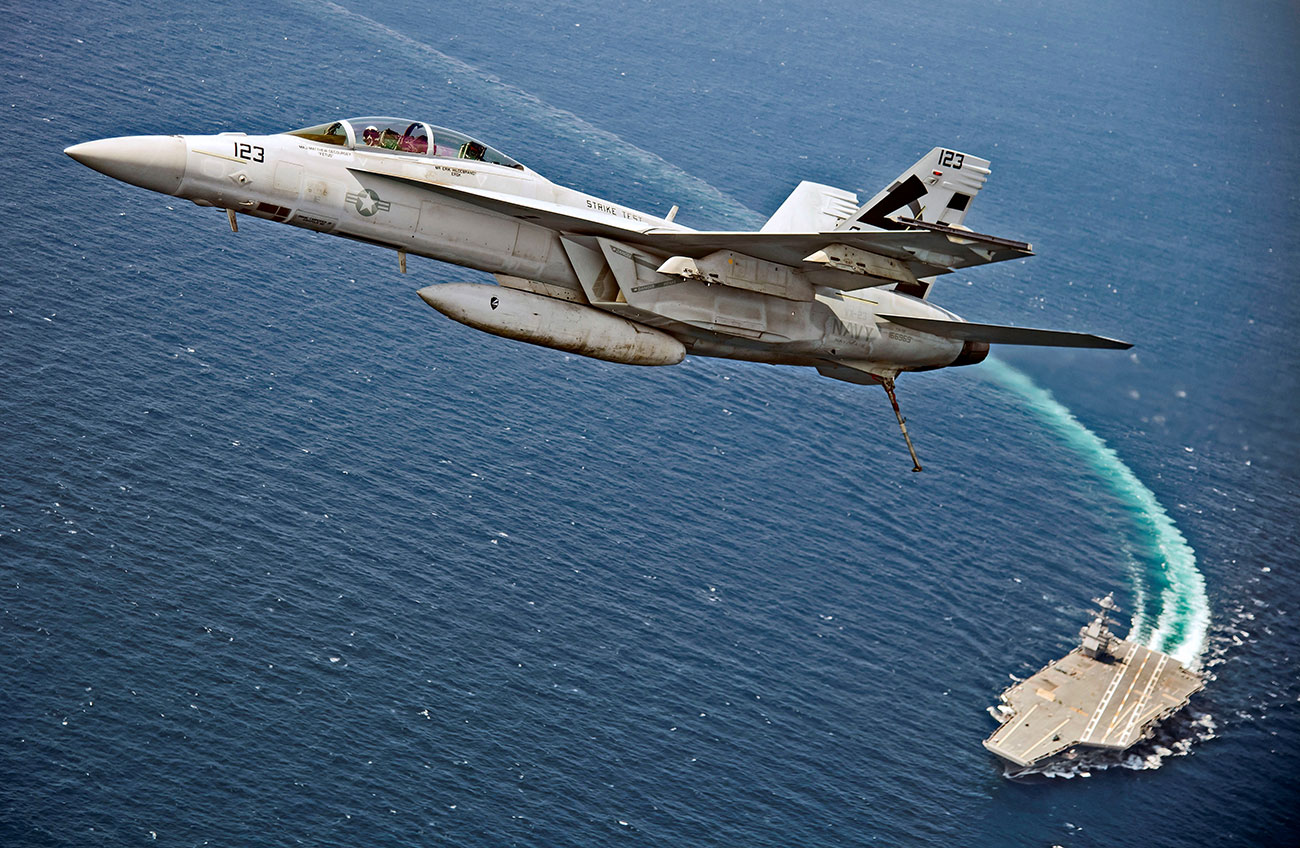 An F/A-18F Super Hornet jet flies over the USS Gerald R. Ford as the U.S. Navy aircraft carrier tests its new launch and flight arrest systems