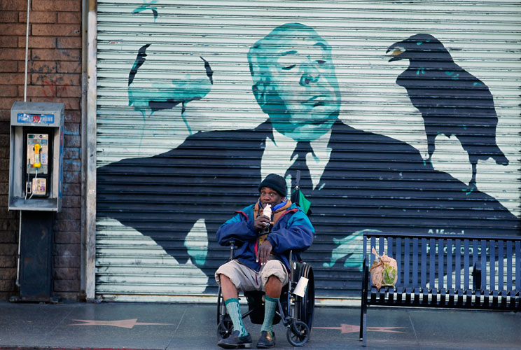 Store front metal doors on Hollywood boulevard depict the image of film director Alfred Hitchcock as a man in a wheelchair sips on a drink.