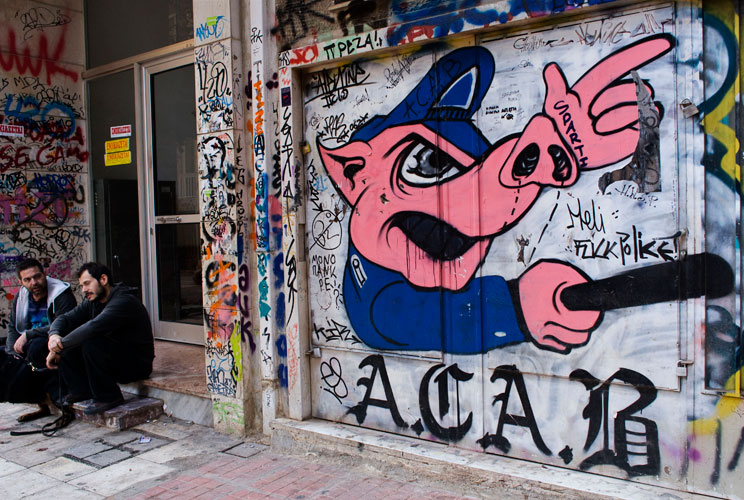 Men sit near graffiti displayed on a building on December 6, 2011 in Athens, Greece. Graffiti artists throughout the city are expressing the effects of austerity measures that have plagued the community as Greece continues to struggle in debt while lawmak