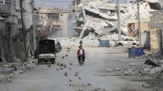 Birds fly near men riding a motorcycle through a damaged neighbourhood in the northern Syrian town of al-Bab