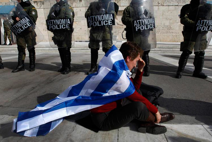 An anti-austerity protester draped with a Greek flag sits in front of police guarding parliament in Athens February 11, 2012 during a demonstration on the second day of a 48-hour strike by Greek workers unions.